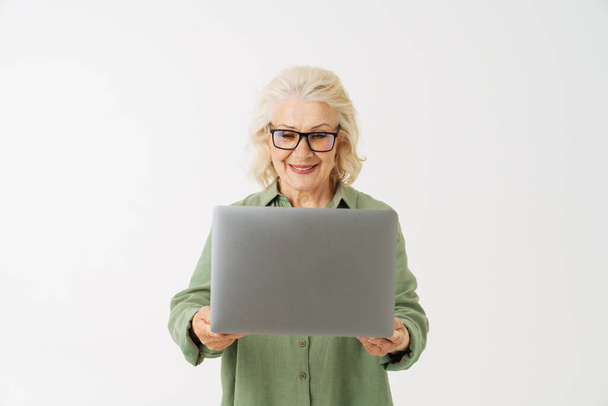 Grey senior woman in eyeglasses smiling while posing with laptop isolated over white background - Fotoğraf, Görsel
