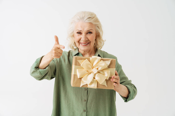 Grey senior woman pointing finger and showing gift box at camera isolated over white background - Fotó, kép