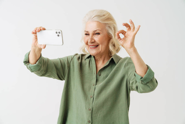 Grey senior woman gesturing while taking selfie on mobile phone isolated over white background - Foto, afbeelding