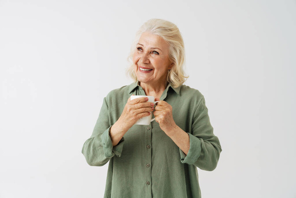 Grey senior woman in shirt smiling and drinking coffee isolated over white background - Foto, Imagem