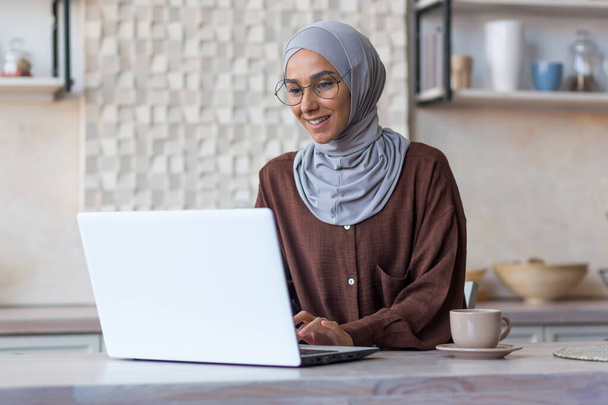 Młoda piękna muzułmanka w hidżabie i okulary studiuje zdalnie przy użyciu laptopa student kobieta w kuchni uśmiechnięta i szczęśliwa. - Zdjęcie, obraz