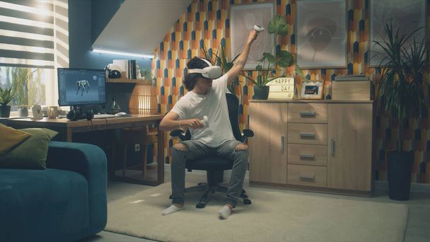 Mann spielt Videospiel mit modernem VR-Headset und drahtlosen Controllern in der Freizeit in seinem Zimmer. Virtual-Reality-Gerät. Cyberspace und Metaverse - Foto, Bild