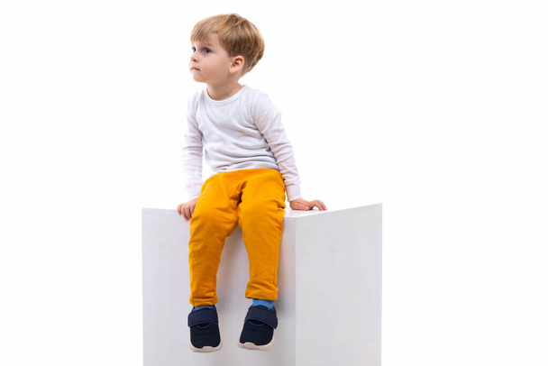 Egy kíváncsi gyerek ül egy fehér kockán egy fehér falon, balra néz. - Fotó, kép