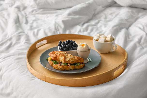 Vassoio di legno con gustoso croissant, uovo sodo, mirtillo e tazza di cacao sul letto - Foto, immagini