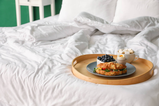 Vassoio in legno con gustoso croissant, uovo sodo, mirtillo e tazza di cacao su un comodo letto - Foto, immagini