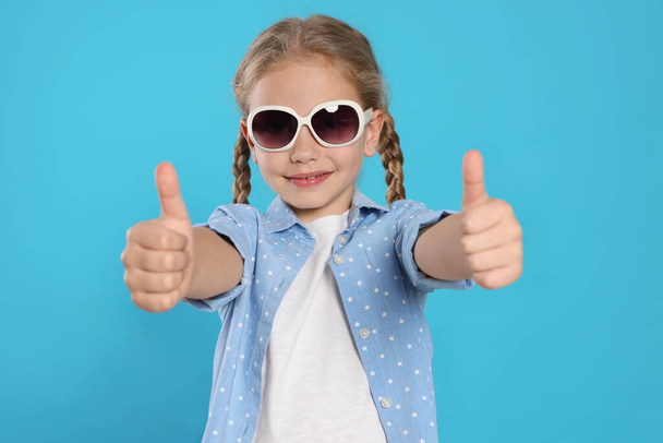 Girl in stylish sunglasses showing thumbs up on light blue background - Foto, afbeelding
