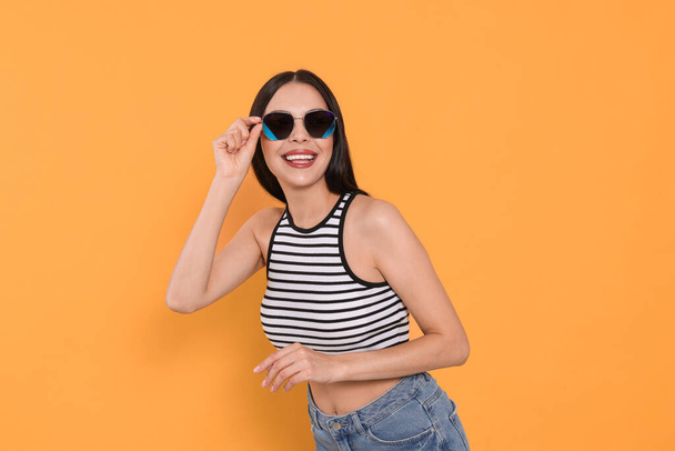 Attractive happy woman touching fashionable sunglasses against orange background - Foto, Imagen