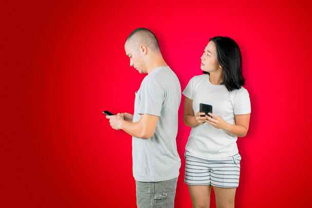 Asian young woman spying at cellphone of her husband while standing together in the studio with red background - 写真・画像
