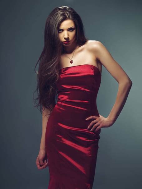 Danger never looked so tempting. Fashion model posing sensually in a fitting red satin dress against a dark background - Photo, Image