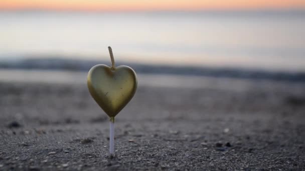 Un homme allume une bougie en forme de cœur sur la plage de sable de la mer au coucher du soleil de l'aube. Bougie en forme de coeur brûlant sur fond de vagues de mer à l'aube gros plan. Ambiance romantique. Saint Valentin - Séquence, vidéo