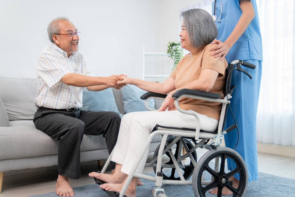 Ένα ικανοποιημένο ζευγάρι ηλικιωμένων και η νοσοκόμα τους στο σπίτι. Ηλικιωμένη γυναίκα σε αναπηρική καρέκλα με το νεαρό φροντιστή της. - Φωτογραφία, εικόνα