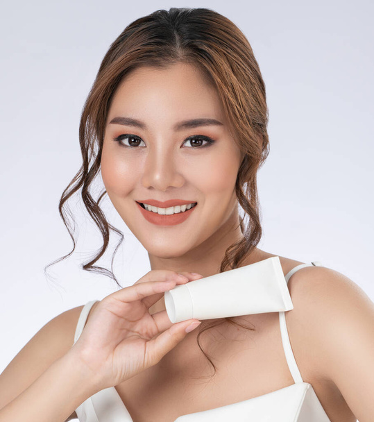 Gorgeous woman with makeup smiling holding mockup product for advertising text place, light grey background. Advertising concept for skin healthcare and beauty care products. - Photo, Image
