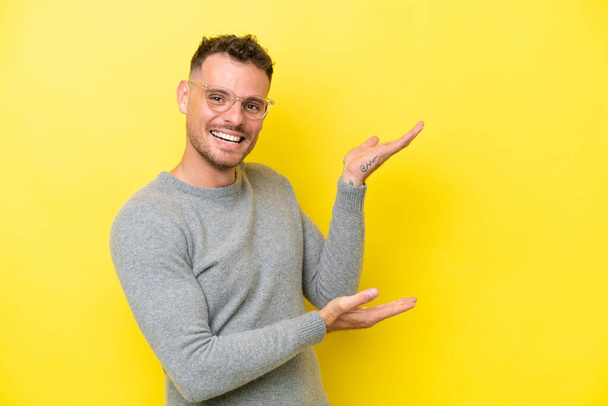 Young caucasian handsome man isolated on yellow background extending hands to the side for inviting to come - Fotó, kép