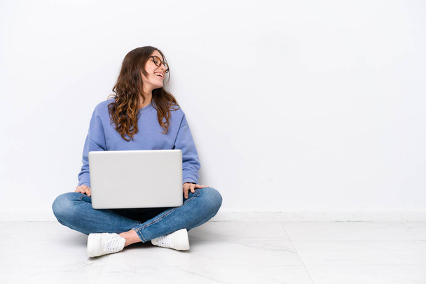Yerde dizüstü bilgisayarlı beyaz bir kadın oturuyor. Beyaz arka planda izole edilmiş. Yan pozisyonda gülüyor. - Fotoğraf, Görsel