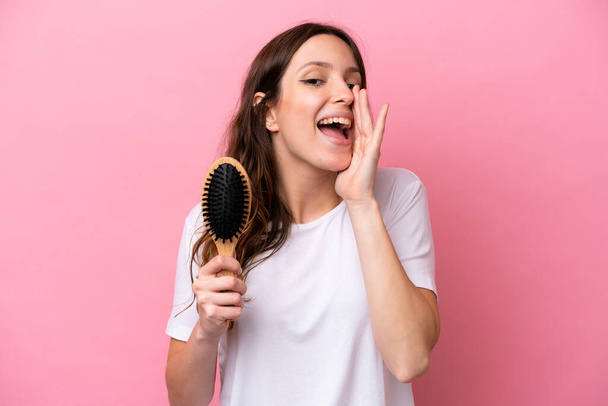 Young caucasian woman with hair comb isolated on pink background shouting with mouth wide open - Фото, изображение