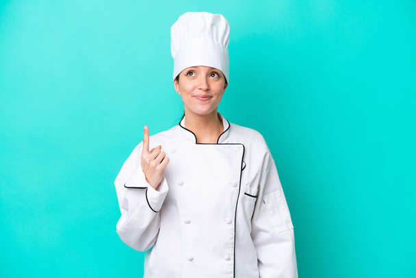 Молодая белая шеф-повар женщина изолирована на синем фоне указывая указательным пальцем отличная идея - Фото, изображение