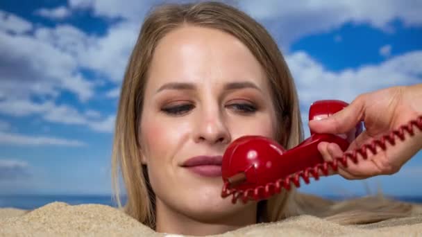 Красивая женщина, закопанная по самую голову на пляже, разговаривает по красному винтажному телефону - Кадры, видео