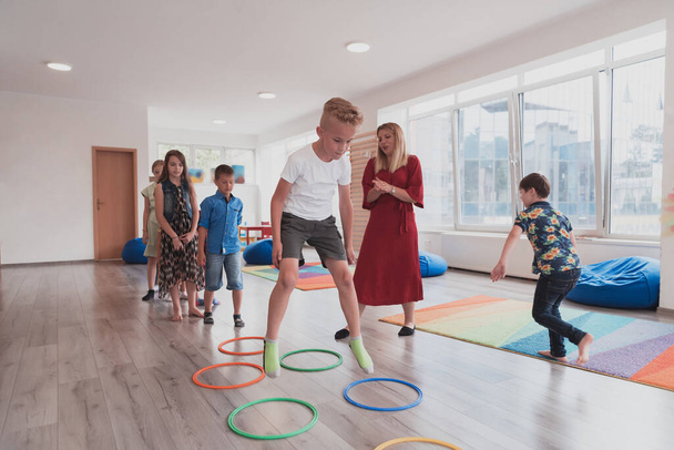 Small nursery school children with a female teacher on the floor indoors in the classroom, doing exercise. Jumping over hula hoop circles track on the floor. High quality photo - Foto, Imagem