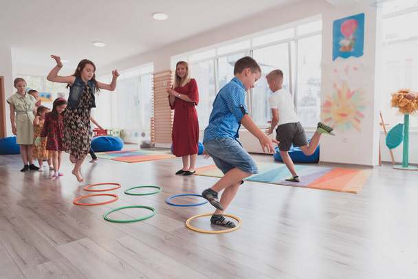 Small nursery school children with a female teacher on the floor indoors in the classroom, doing exercise. Jumping over hula hoop circles track on the floor. High quality photo - Valokuva, kuva