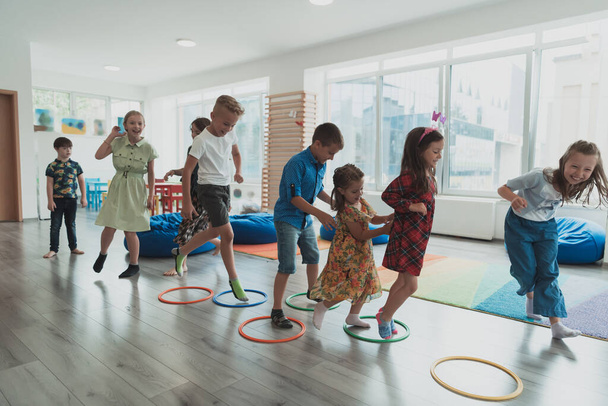 Small nursery school children with a female teacher on the floor indoors in the classroom, doing exercise. Jumping over hula hoop circles track on the floor. High quality photo - Photo, Image