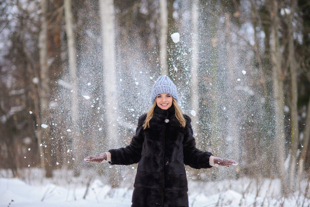 portrait d'une belle blonde souriante en manteau de fourrure noir et chapeau bleu sur fond de forêt enneigée hivernale. Elle jette de la neige et elle vole dans les airs - Photo, image