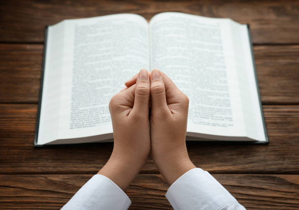 Woman holding hands clasped while praying at wooden table with Bible, closeup - Foto, afbeelding