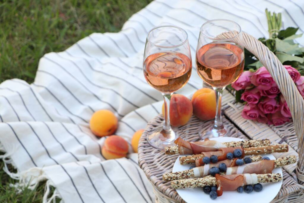 Glasses of delicious rose wine, food, flowers and basket on picnic blanket outdoors - 写真・画像