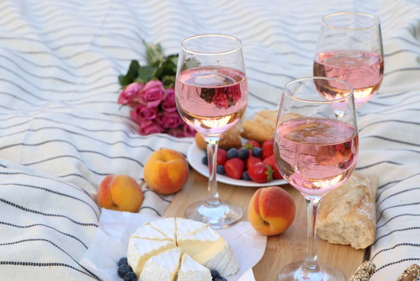Glasses of delicious rose wine, flowers and food on white picnic blanket - 写真・画像