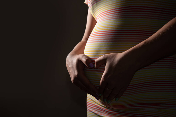 Девушка обнимает свой беременный живот, складывая руки, как сердце. Она в платье на темном фоне студии - Фото, изображение