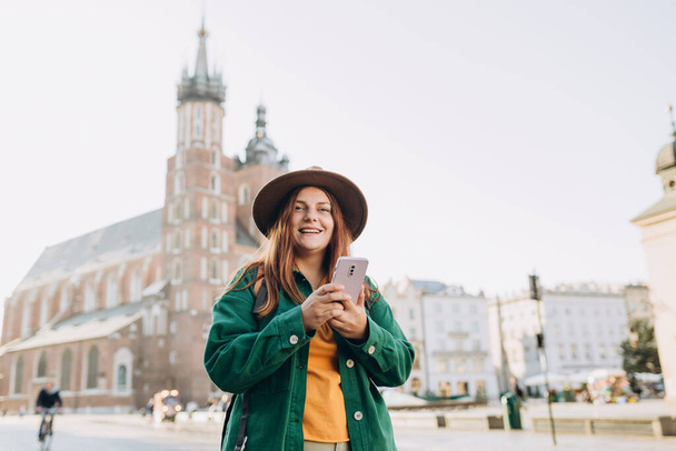Beautiful stylish woman walking on Market Square in Krakow on autumn day and holding mobile phone. Phone Communication. Urban lifestyle concept. Check social networks - 写真・画像