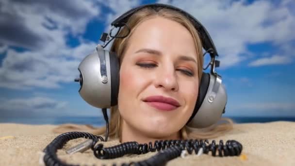 Una mujer bonita enterrada en su cabeza en una playa escuchando música con auriculares - Metraje, vídeo