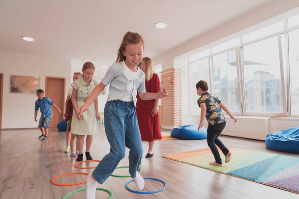 Small nursery school children with a female teacher on the floor indoors in the classroom, doing exercise. Jumping over hula hoop circles track on the floor. High quality photo - Fotó, kép