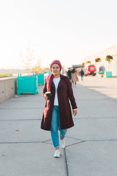 Mulher bonita árabe jovem em hijab andando na rua, tocando no smartphone e beber café para ir. Mulher muito muçulmana em lenço de cabeça passeando no aeroporto, mensagem de texto e bebida sorvendo. - Foto, Imagem