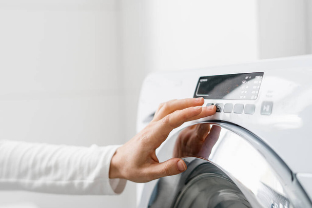 tiro recortado da mão da mulher ligue a máquina de lavar roupa automática ou selecione o programa com botão no painel de controle no banheiro branco na casa moderna - Foto, Imagem