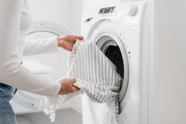 oříznutý záběr ženských rukou vložte ložní prádlo nebo bavlněný ručník do automatické pračky s otevřenými dveřmi v koupelně, domácí práce - Fotografie, Obrázek