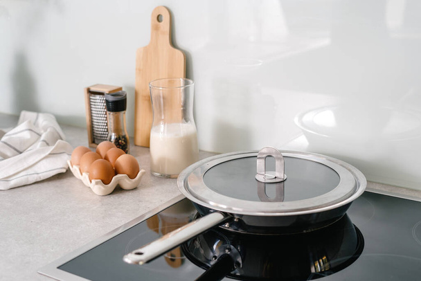 patelnia ze stali nierdzewnej z pokrywą na szklanej ceramicznej płycie kuchennej, jajka, mleko, pieprz i ręcznik na blat w kuchni z jasnym wnętrzem, przygotowując śniadanie w domu - Zdjęcie, obraz
