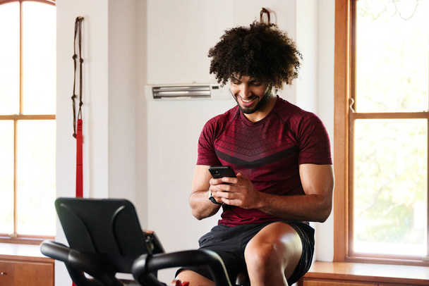 Happy young middle eastern man in sportswear using mobile phone on exercise bike at the gym - 写真・画像