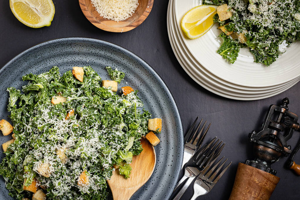 La salade Kale Cesar servait de style familial dans un bol avec une pile de assiettes blanches sur le côté. Moulin au poivre noir, tranche de citron et parmesan râpé comme accessoires. - Photo, image