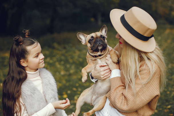 Woman, little girl and dog play and have fun in the autumn park. Brown french bulldog with female owners spend a day at the park playing and having fun. Woman wearing beige coat and a hat. - Photo, Image