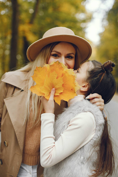 Woman and her little girl walking in the autumn park. Mother and daughter posing for a photo with an autumn leaves. Woman wearing beige coat and a hat. - Foto, immagini