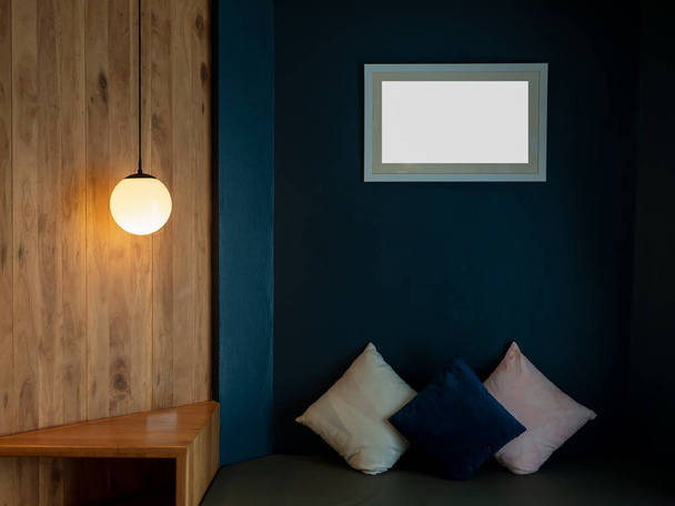 Empty white blank picture frame mockup hanging on blue wall background over the bench and pillows in the dark living room decorated with round ceiling lamp, full moon shape on wood wall. - Fotó, kép