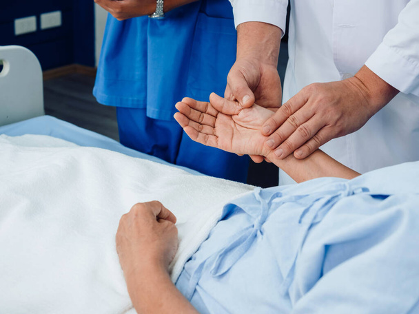 Doctor's hand in white lab coat is holding elderly patient's hand lying on the bed for check to examine heartbeat pulse by fingers in recovering room in hospital. Medical health care checking concept. - Photo, image