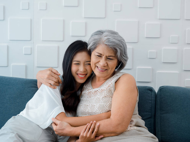 Портрет счастливого азиатского старшеклассника, матери или дедушки с седыми волосами, обнимающих ее прекрасную дочь или внука, улыбающихся с любовью, заботой и комфортом, сидя на сером диване в гостиной дома. - Фото, изображение