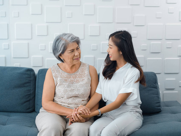 Retrato de feliz Asiático senior, madre o abuelo pelo blanco cogido de la mano de su hermosa hija o nieto sonriendo con amor, cuidado y comodidad mientras se sienta en el sofá gris en la sala de estar. - Foto, imagen