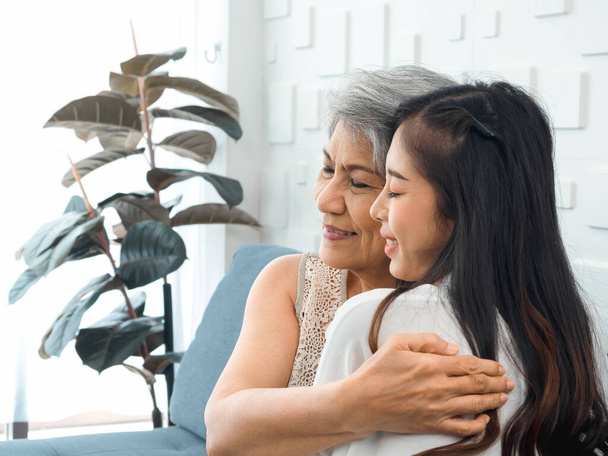 Close-up portrait of happy Asian senior, mother or grandparent white hair embracing her beautiful daughter or grandchild with love, care and comfort while sit on grey couch in living room at home. - Photo, Image