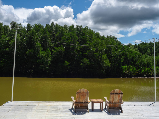 Dos sillas de madera vacías solitarias y una mesa auxiliar en la terraza de madera decorada con iluminación festiva de río y vista verde de la selva tropical en un día soleado. Relax y lugar de paz en el campo. - Foto, Imagen