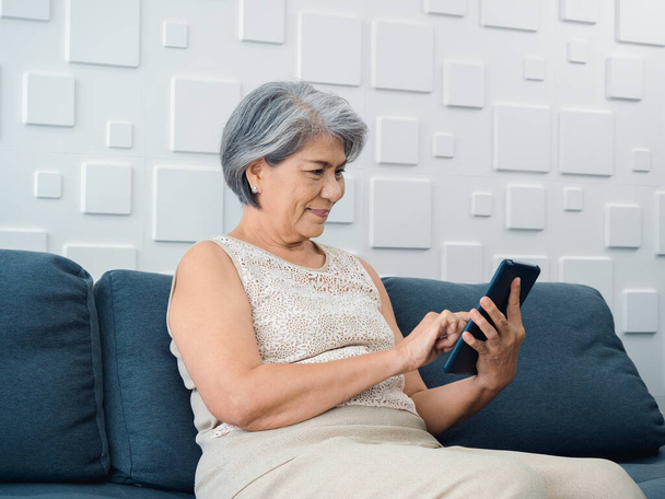 Szczęśliwa azjatycka starsza kobieta siedzi na kanapie, trzymając i patrząc na cyfrowy ekran tabletu w ręce w salonie. Starsza kobieta używająca inteligentnego urządzenia, komputer w domu. Starsi ludzie używają technologii z łatwością. - Zdjęcie, obraz