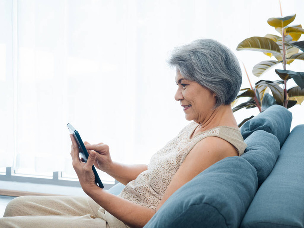 Счастливая азиатская старшая женщина сидит на диване, держа и глядя на цифровой экран планшета в руке в гостиной. Пожилая женщина использует смарт-устройство, компьютер дома. Пожилые люди используют технологию с легкостью. - Фото, изображение