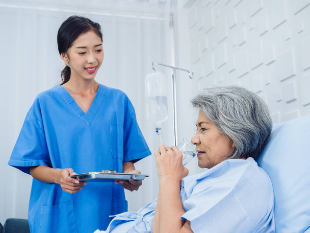 Asian elderly patient taking medicine, hold drinking water glass and pill from young nurse in blue scrub suit in hospital room, daily medicine or vitamin supplements, senior healthcare and medical. - Photo, Image