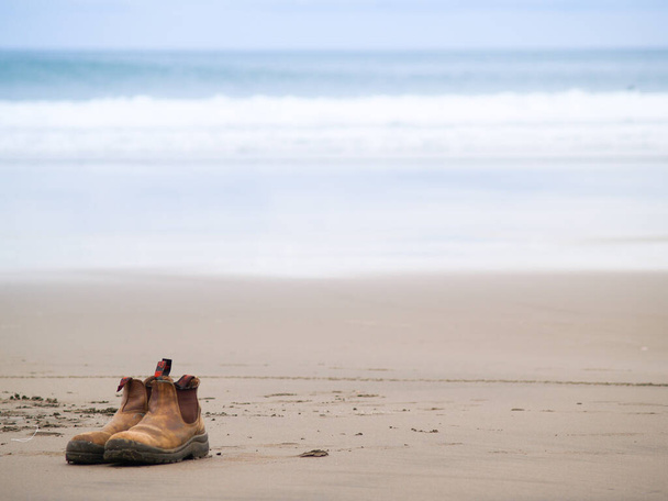 Par de botas de cuero viejo en la arena en la playa con la impresión de la playa y el surf. - Foto, imagen